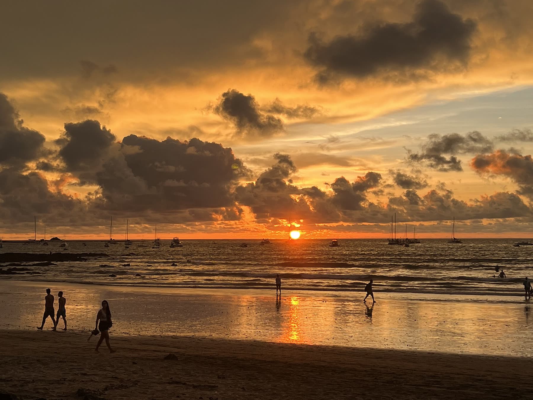 People strolling along a Pacific beach as the sun sets behind golden clouds